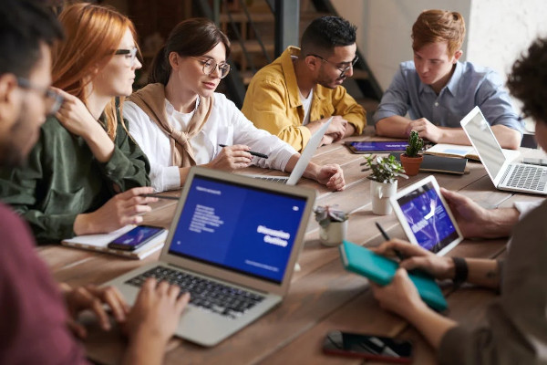 Eine Gruppe junger Menschen sitzt mit ihren Laptops an einem Holztisch und unterhalten sich